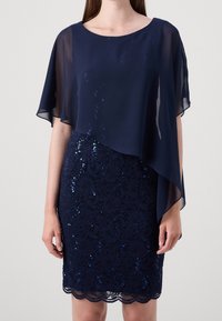 Žena v tmavě modré šaty s flitry a krajkovým detailem, s asymetrickým průhledným pláštěm a krátkými rukávy.