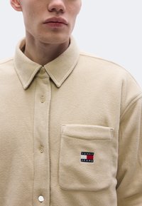 Bézs színű polár ingdzseki galléros nyakkal, gombos záródással és elülső zsebbel, amelyen "Tommy Jeans" logó hímzés található. Texturált, puha anyagból.