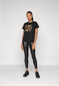 T-shirt nera con grafica a stampa leopardata, abbinata a leggings neri lucidi. Modella che indossa scarpe da ginnastica nere, in piedi su uno sfondo neutro.