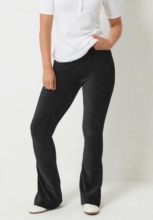 Persoon die zwarte flared corduroy broek draagt, wit kortermig overhemd en witte schoenen, staand met één hand in de zak.