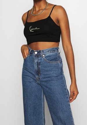 Top court noir avec détail signature vert, associé à un jean large taille haute en denim bleu clair. Bretelles courtes, texture lisse.