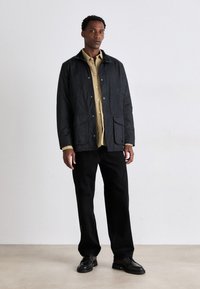 Veste légère noire avec deux poches avant, superposée sur une chemise beige à boutons. Le modèle porte un pantalon noir et des chaussures noires.
