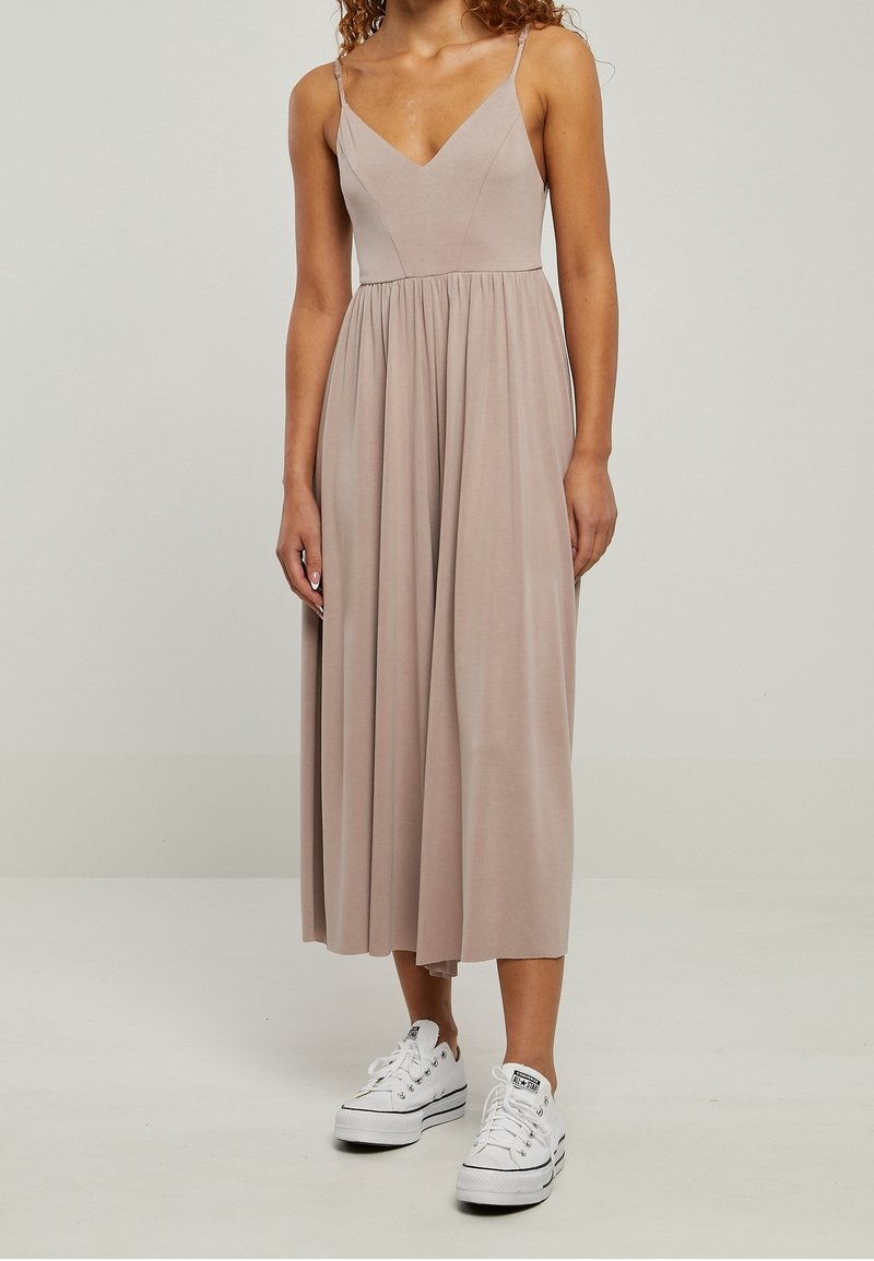 Femme portant une robe midi beige sans manches avec un col en V et des baskets blanches à plateforme, debout devant un fond uni.