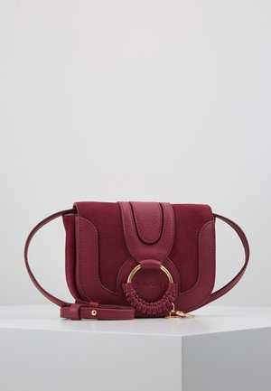 Pequeño bolso bandolera burdeos de cuero y ante con detalle de anillo dorado trenzado y correa ajustable sobre una superficie blanca.