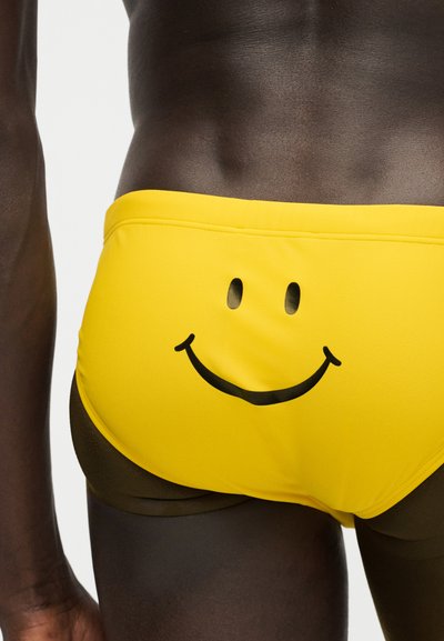 Κοντινό πλάνο ενός ατόμου που φοράει κίτρινα μαγιό σλιπ με σχέδιο μαύρου χαμογελαστού προσώπου στο πίσω μέρος.