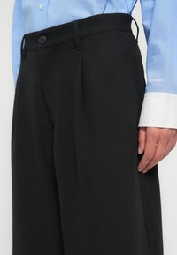 Μαύρο παντελόνι ραμμένο στα μέτρα με ψηλή μέση, πιέτες μπροστά και πλαϊνές τσέπες. Το ύφασμα φαίνεται λείο, σε συνδυασμό με ένα ανοιχτό μπλε πουκάμισο.