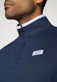 Pullover azul-marinho com ziper de um quarto e colarinho branco, apresentando um rótulo da marca. Material suave com textura aveludada. Vista aproximada da área do pescoço.