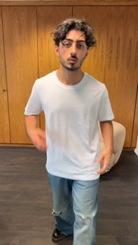 Un tricou alb din bumbac, cu mâneci scurte, asortat cu blugi deschiși, cu un aspect uzat, și pantofi negri, stând pe un podea din lemn închis la culoare.