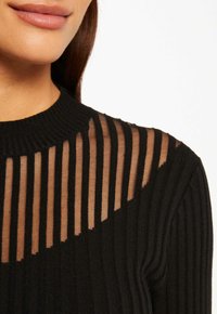 Pull noir côtelé avec un motif de rayures transparentes sur le haut de la poitrine. L'encolure est côtelée, offrant un contraste texturé.