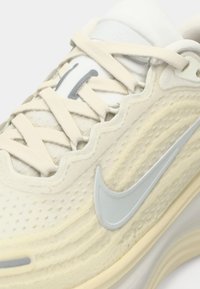 Κοντινό πλάνο ενός μπεζ και λευκού αθλητικού παπουτσιού Nike με κυματιστό σχέδιο, υφασμάτινη υφή και ανοιχτό γκρι λογότυπο swoosh στο πλάι.