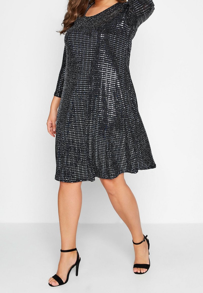 Kvinna klädd i en svart och silver paljettklänning som når till knäna, med trekvarts ärm och svarta högklackade sandaler, poserar.