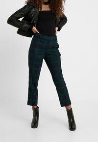 Vihreät ja laivastonsiniset ruutukuvioiset housut, joissa on räätälöity istuvuus, sivutaskut ja lyhyt lahkeen pituus. Yhdistetty istuvaan mustaan toppiin ja nahkatakkiin.