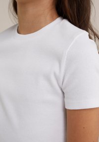 Camiseta corta de manga corta blanca con un clásico cuello redondo. La tela parece suave y elástica con una textura lisa.
