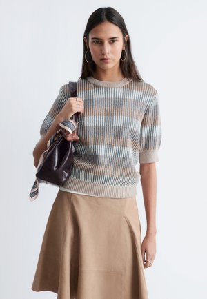 Kvinde med langt mørkt hår iført en kortærmet sweater med blå, beige og hvide striber, tan-farvet nederdel og holder en mørk skindtaske med et tørklæde.