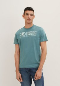 Teal bomulds T-shirt med korte ærmer, rund halsudskæring og hvid grafisk tekst på forsiden. Båret med blå jeans.
