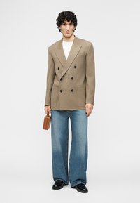 Άνδρας που φοράει μπεζ σακάκι διπλής επίστρωσης, λευκό πουκάμισο, μπλε τζιν με φαρδιά πόδια, μαύρα παπούτσια, κρατώντας μικρή καφέ τσάντα σε απλό φόντο.