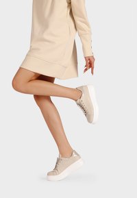 Sneakers in pelle beige con finitura strutturata, punta rotonda, accenti argentati e suola spessa bianca; esposti accanto a un vestito beige.