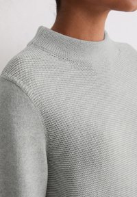 Jasnoszary sweter z dzianiny z wysokim kołnierzem, z fakturowanym wzorem i prążkowymi wykończeniami, ukazujący szczegóły tkaniny i bezszwowe ramiona.