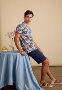 Bloemige shirt met korte mouwen in crèmekleurige, blauwe en oranje tinten, gecombineerd met marineblauwe shorts; model leunt tegen een lichtblauwe tafelkleed.