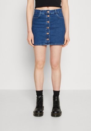 Femme portant une mini-jupe en denim bleue à boutons devant, un top court noir, des chaussettes noires et des bottes noires lacées, debout devant un fond uni.