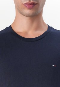 Close-up van het onderste deel van het gezicht en de nek van een persoon die een donker marineblauw shirt met ronde hals draagt met een klein rood-wit-blauw logo op de borst.