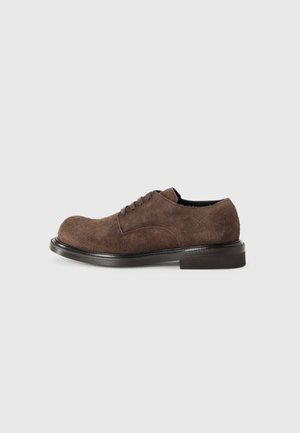 Brown suede lace-up shoe with a rounded toe, flat black sole, and minimalistic design. The texture appears soft and slightly fuzzy.
