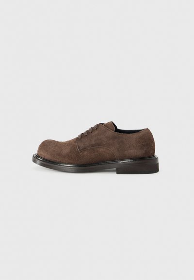 Hnědé semišové šněrovací boty s zaoblenou špičkou, plochou černou podrážkou a minimalistickým designem. Textura vypadá měkce a mírně jemně.