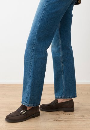 Personne portant un jean bleu droit et des mocassins en daim brun foncé avec une boucle sur un sol en bois.