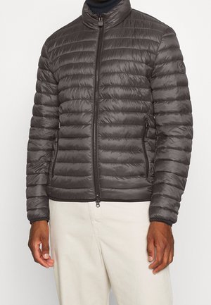 Mężczyzna ubrany w ciemnoszary pikowany kurtkę na suwak ze stójką oraz jasnobeżowe spodnie na jednolitym tle.