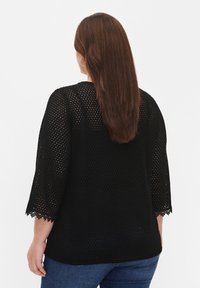 Femme aux longs cheveux bruns portant des boucles d'oreilles créoles, un haut noir perforé avec bordure en dentelle, et un jean bleu, vue de dos sur fond blanc.