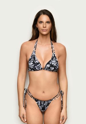 Mujer con cabello largo y castaño, que lleva un bikini con estampado floral en blanco y negro, de pie frente a un fondo claro y simple.
