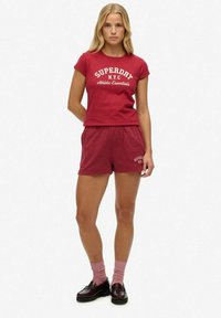 T-shirt en coton rouge et shorts assortis, avec le texte blanc "SUPERDAY NYC Athletic Essentials". Stylé avec des chaussettes roses et des chaussures noires.