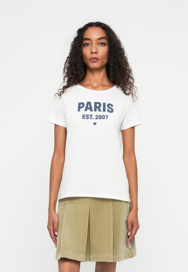 White t-shirt with navy text "PARIS EST. 2007" and a small heart icon, paired with a pleated, light olive green corduroy skirt.