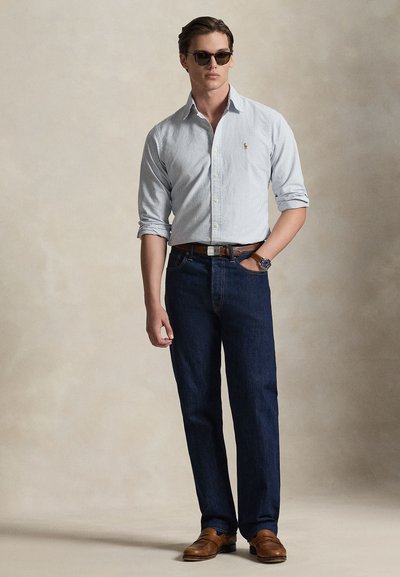 Világoskék csíkos gumgombos ing, sötétkék farmer, barna bőr öv és cipők, semleges háttér előtt. Kiegészítők között napszemüveg is van.