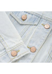 Ljusblå denimjacka med guldstickning och kopparfärgade knappar med en solstrålesdesign. Detaljerad ficklock synlig.