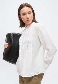 Kvinne iført hvit bluse og beige bukser holder en stor, svart rektangulær pute mot hvit bakgrunn.