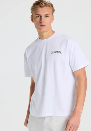 Ung mand med kort blondt hår iført en hvid t-shirt med "CASPARA" tekst på brystet og lysegrå bukser, stående mod en hvid baggrund.