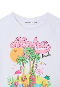 T-shirt bianca in cotone con un design stampato colorato, arricchito da rhinestone rosa, palme, fenicotteri e tavole da surf su uno sfondo da spiaggia.
