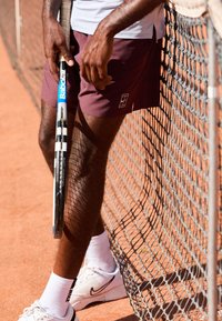 Tennis lühikesed püksid tumepruunis, logo olemas. Tennisereket siniste ja mustade aktsentidega, toetatud tenniseteeni saviväljakul.