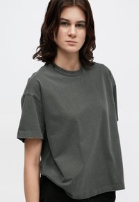 Pelēks kokvilnas T-krekls ar apaļu kakla izgriezumu un īsām piedurknēm, kas raksturo brīvu griezumu un minimālu šuvju detaļu.