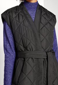 Svart kviltad väst med bred axeldesign, bältesmidja och diagonala sömnadsdetaljer, bärs över en strukturerad lila turtleneck.