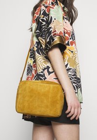 Mujer vestida con una camisa de flores coloridas de manga corta y pantalones cortos negros, llevando un bolso cruzado de ante en color mostaza.