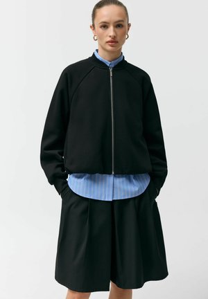 Kobieta w czarnej kurtce na zamek błyskawiczny, na niebieskiej koszuli w paski i czarnych szerokich szortach, stojąca z rękami w kieszeniach na białym tle.