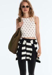 Kobieta w okularach przeciwsłonecznych, białym, bezrękawnym topie w kropki, czarno-białym swetrze w paski związanym w talii, czarnych spodniach i z brązową torbą.