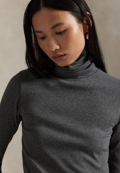 Noor nais pika tumeda juustega vaatab alla, kandes kehale järgivat tumedat halli soonikkoondavat kõrge kaelusega kampsunit ja väikseid kõrvarõngaid ühevärvilise taustaga.