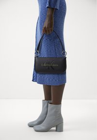 Crna torba Calvin Klein Jeans s utisnutim logotipom, srebrnim hardverom i remenom za rame, u paru s svijetlosivim čizmama na petu do gležnja.