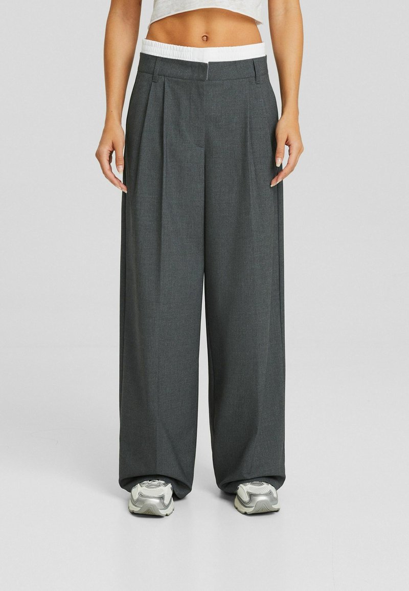 Pantalons à jambes larges en tissu gris foncé, dotés d'une ceinture élastique blanche, de plis à l'avant et de deux poches latérales.