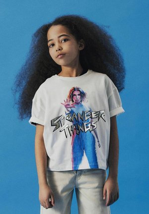 T-shirt blanc avec un graphique d'un personnage de "Stranger Things", associé à un jean bleu et des manches retroussées. L'arrière-plan est d'un bleu uni.