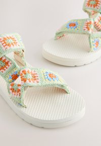 Sandales blanches texturées avec une bride multicolore en crochet doux présentant des motifs floraux pastel verts, roses, orange et bleus.