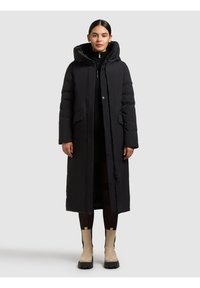 Lang sort isoleret frakke med hætte, med knaplukning og sidelommer, båret over et sort outfit og beige ankelstøvler.